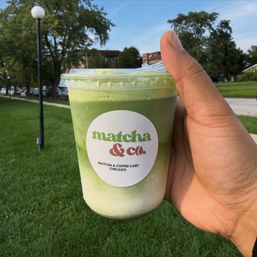 A matcha&co. cup of iced matcha, labelled with the logo, held up in a park